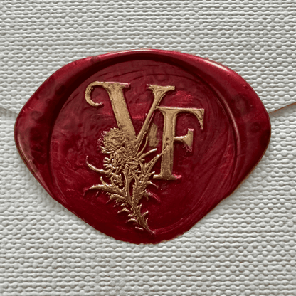 A deep red wax seal embossed with the gold initials "VF" intertwined with a detailed thistle motif. The background is textured white paper, evoking a sense of elegance and historical romance.