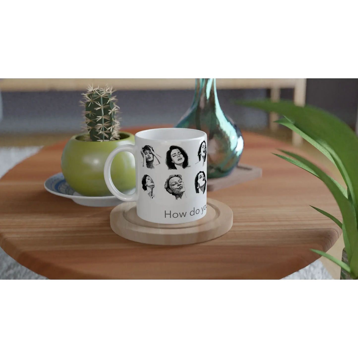 Smut reader mug with expressive black and white illustrated faces, photographed on a wooden table with a cactus and decorative glass vase in a relaxed home setting.
